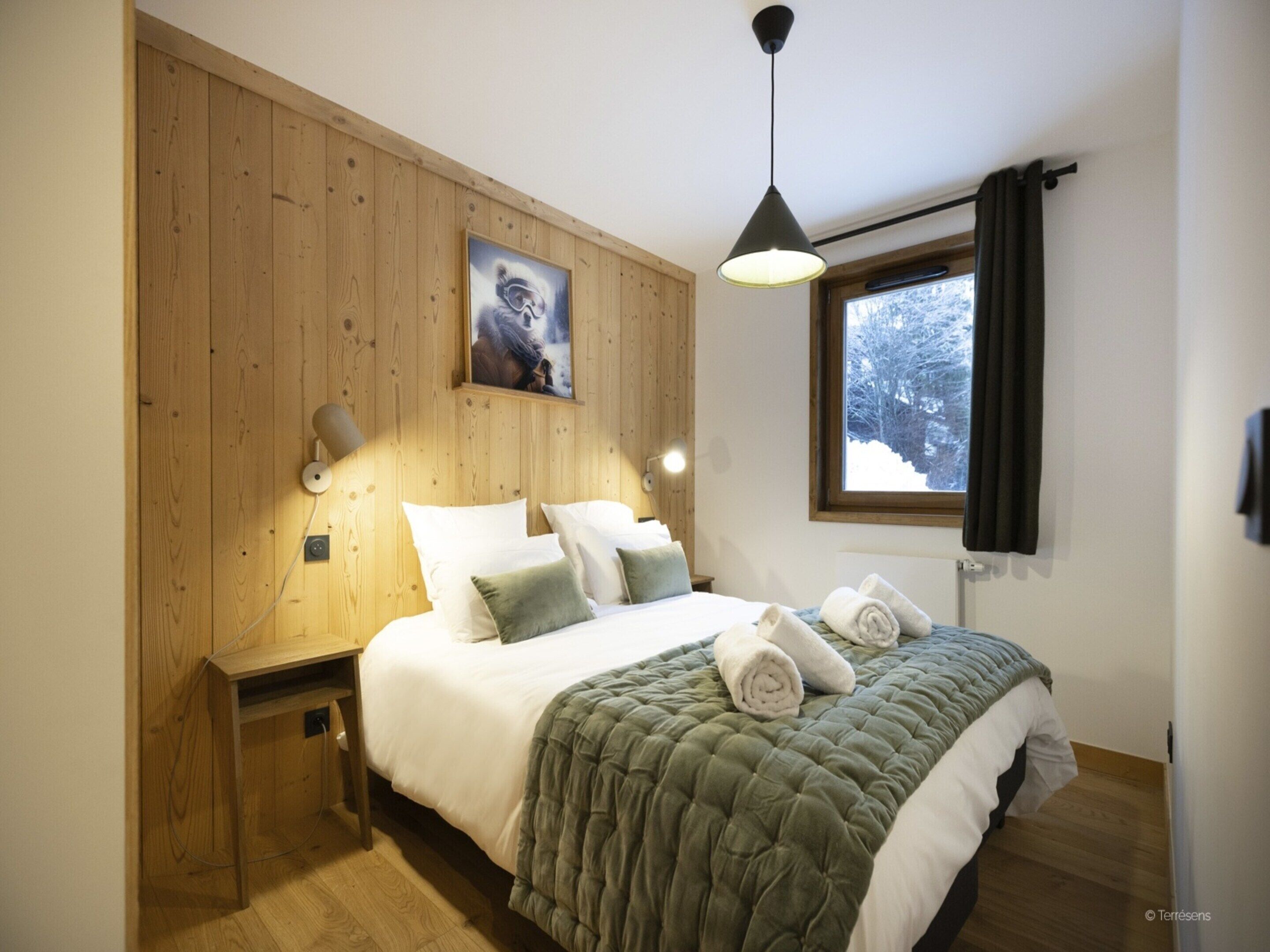 Ein Schlafzimmer mit einer Holzwand, einem Bett mit weißer Bettwäsche und einer grünen Steudecke, einem Fenster mit Blick auf schneebedeckte Bäume und einem Gemälde eines Pferdes an der Wand.