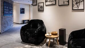 A modern lobby with a stone wall, black velvet chairs, a wooden stool with a cup and bowl, and framed black and white photographs on the wall.
