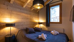 A cozy bedroom with a wooden ceiling, a large bed with dark bedding, two bedside lamps, and a window with a view of a building.