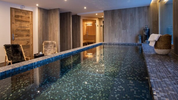 A modern indoor swimming pool with a clear glass bottom, wooden walls, and a seating area with a chair and a basket.