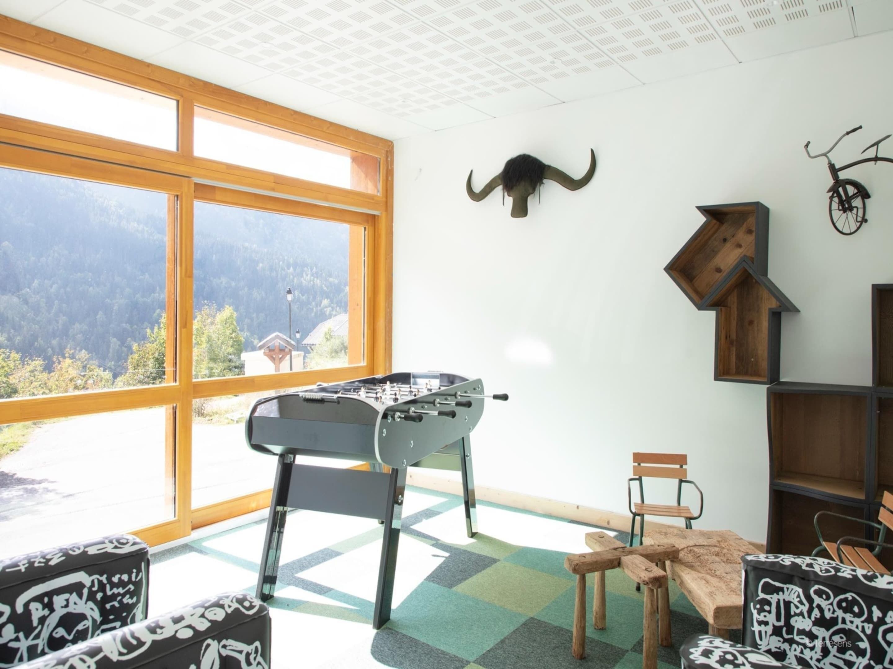 A room with a foosball table, a wooden chair, a sofa, and a mountain view through large windows.
