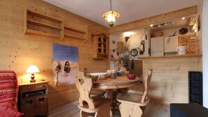 A wooden interior with a round dining table, chairs, a hanging light fixture, and a kitchen area with shelves and appliances. - 1st floor, balcony, tv, 40m², Serre Chevalier (Briançon)