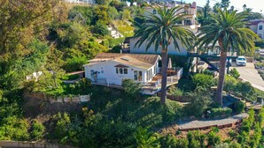 A white house with a balcony, surrounded by lush greenery and palm trees.