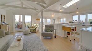 A spacious living area with a sofa, ottoman, and a dining table. There is a kitchen area with a counter and a window overlooking the sea.
