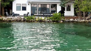 Ein zweistöckiges weißes Haus mit großen Fenstern, einer Steinmauer und einem Steg, der ins Wasser ragt.