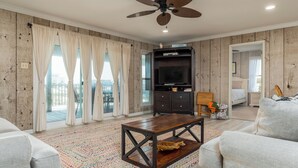 A living room with a wooden floor, a ceiling fan, a TV on a dark wooden cabinet, and a colorful rug. - Canal-front, Boat Dock, Sleeps 9 - Anchor Eways (Dauphin Island)