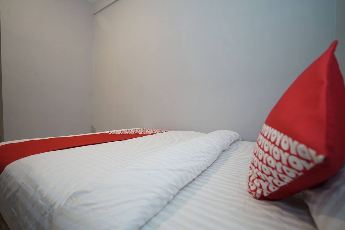 A bed with a white and red bedspread, a red pillow with white text, and a plain white wall.