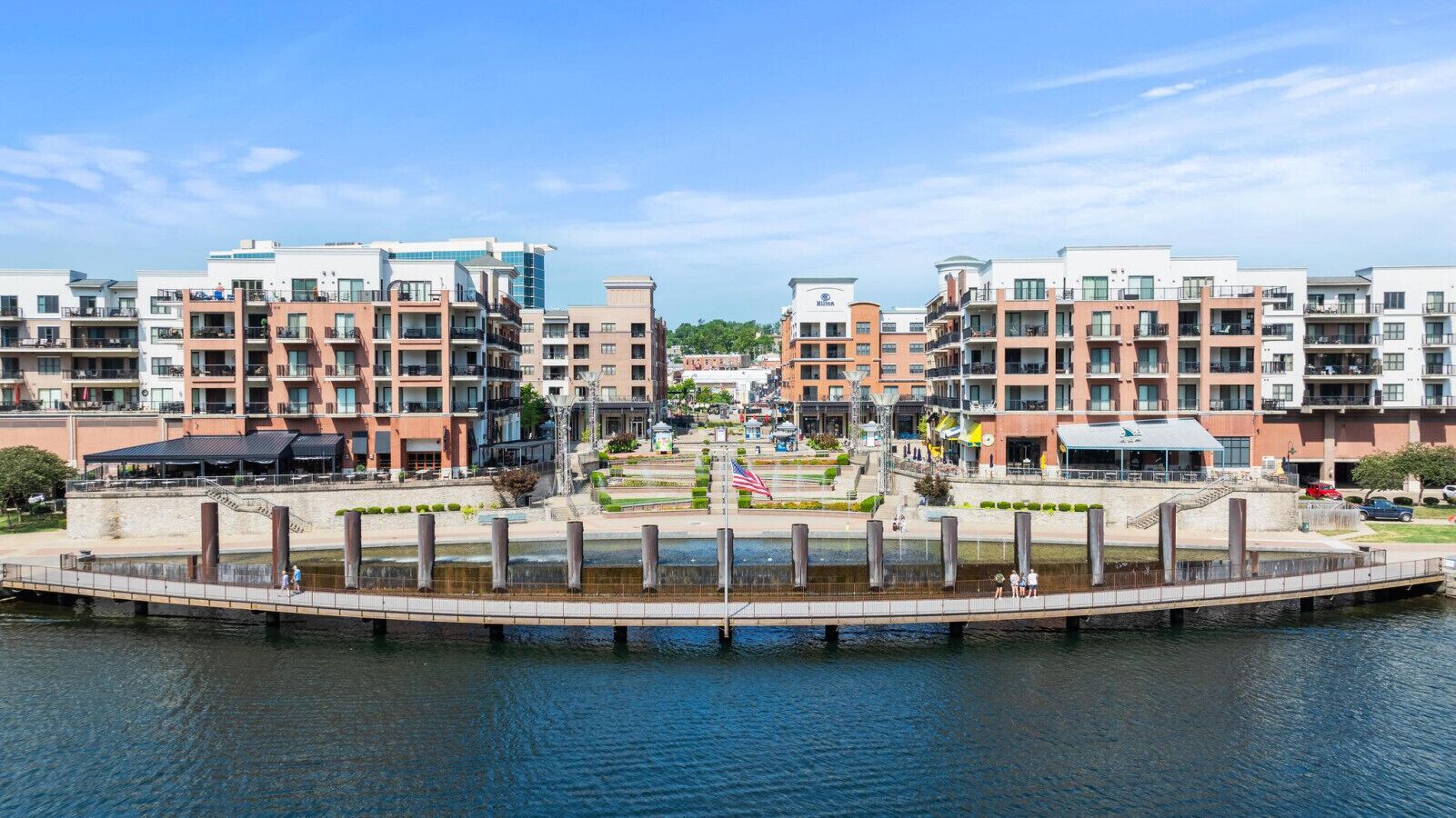 A waterfront hotel with multiple floors, a central courtyard with an American flag, and a pedestrian bridge over the water.
