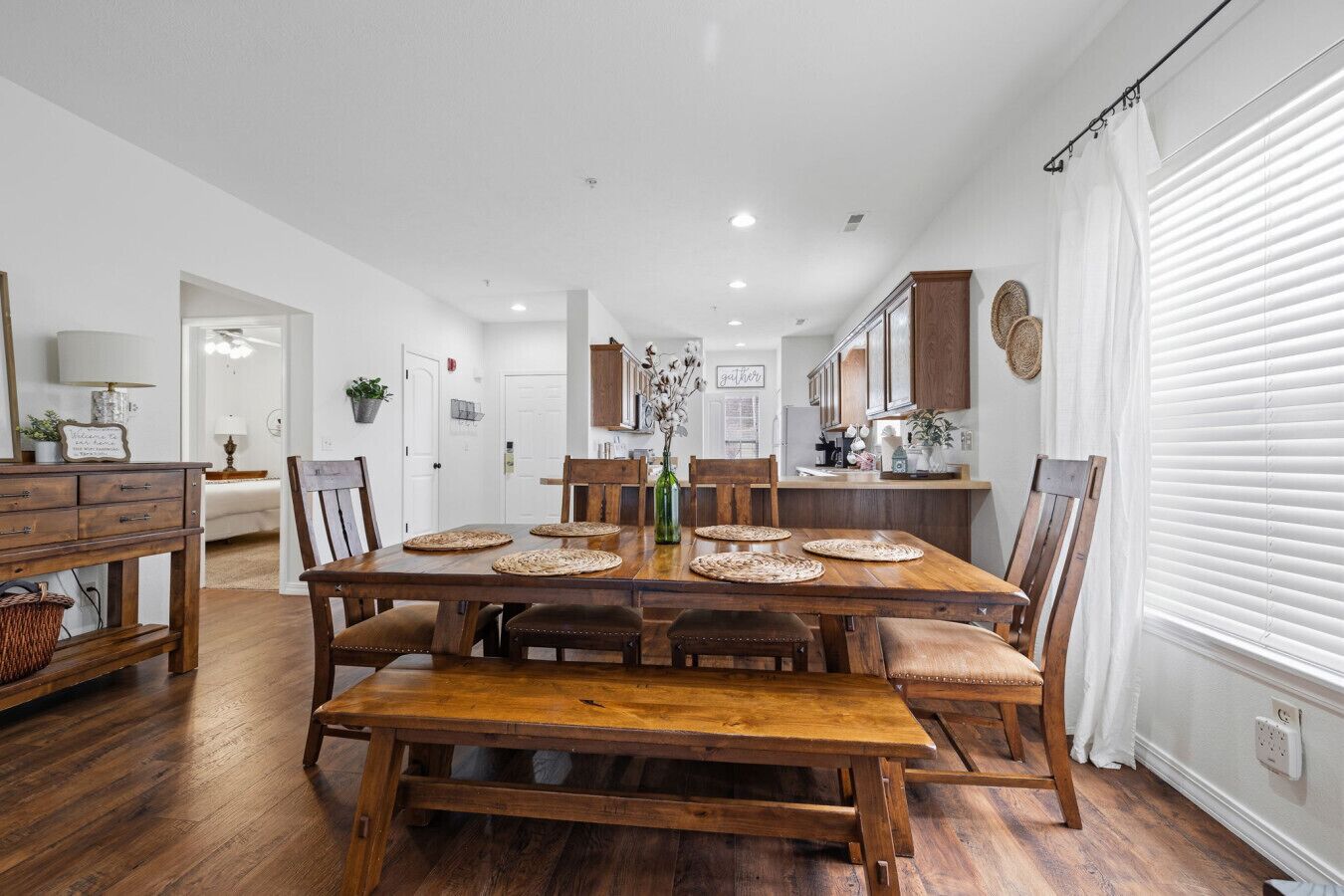 A spacious dining area with a wooden table and benches, a kitchen in the background, and a white interior.