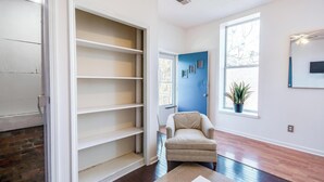 Une pièce avec une grande étagère blanche, un fauteuil beige, une plante en pot près d’une fenêtre et un téléviseur à écran plat fixé au mur.