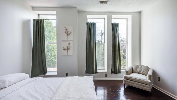 Une chambre à coucher avec un lit, deux fenêtres ornées de rideaux verts, une chaise et un plancher en bois.