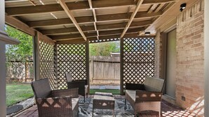 Um pátio externo com mobiliário de vime e um tapete estampado, cercado por uma pérgola de madeira e painéis de treliça.