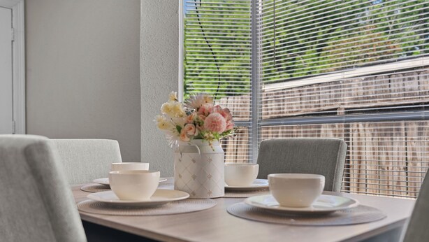 Mesa de jantar com tigelas brancas e um vaso de flores, com uma janela e persianas.