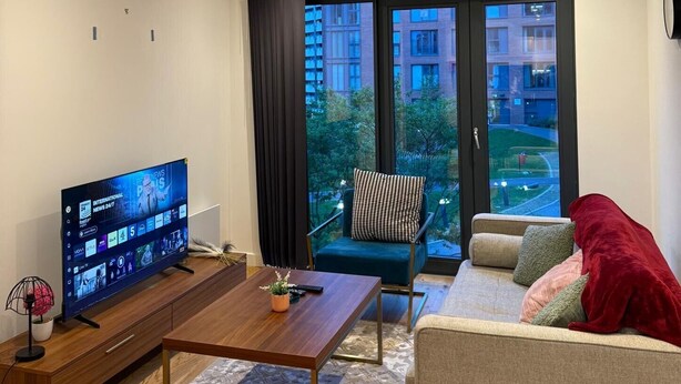 Uma sala de estar moderna com televisão de tela plana, sofá, mesa de centro e vista para uma área residencial através de grandes janelas.