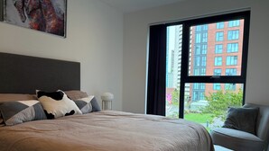 Quarto de hotel moderno com uma cama grande, uma cadeira, uma janela com vista para a cidade e um quadro na parede.