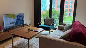 Uma sala de estar moderna com uma TV grande, uma mesa de centro de madeira, uma poltrona azul e um sofá com um cobertor vermelho.
