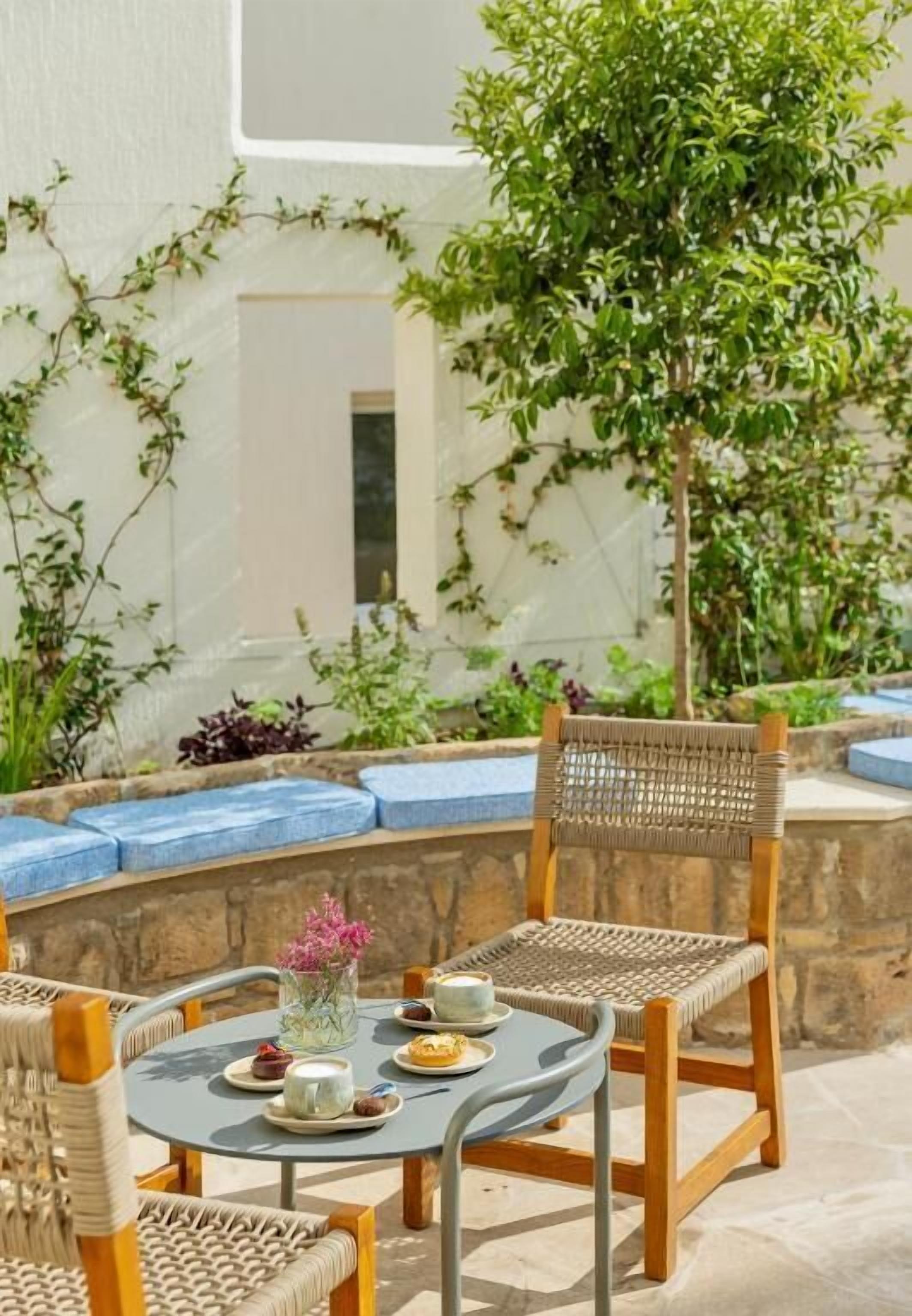 An outdoor seating area with wicker furniture, a round table with a vase of flowers, and a view of a white building and greenery.