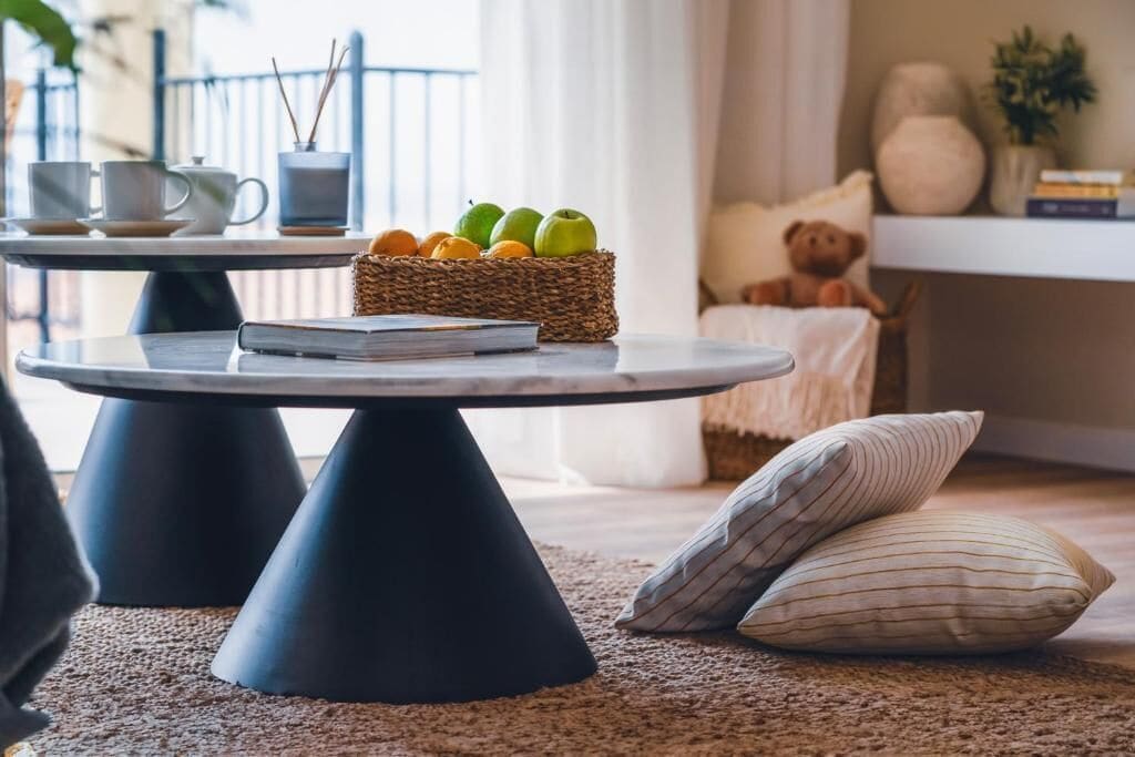 A cozy living room with a round table, a wicker basket with fruits, a teddy bear, and a bookshelf.