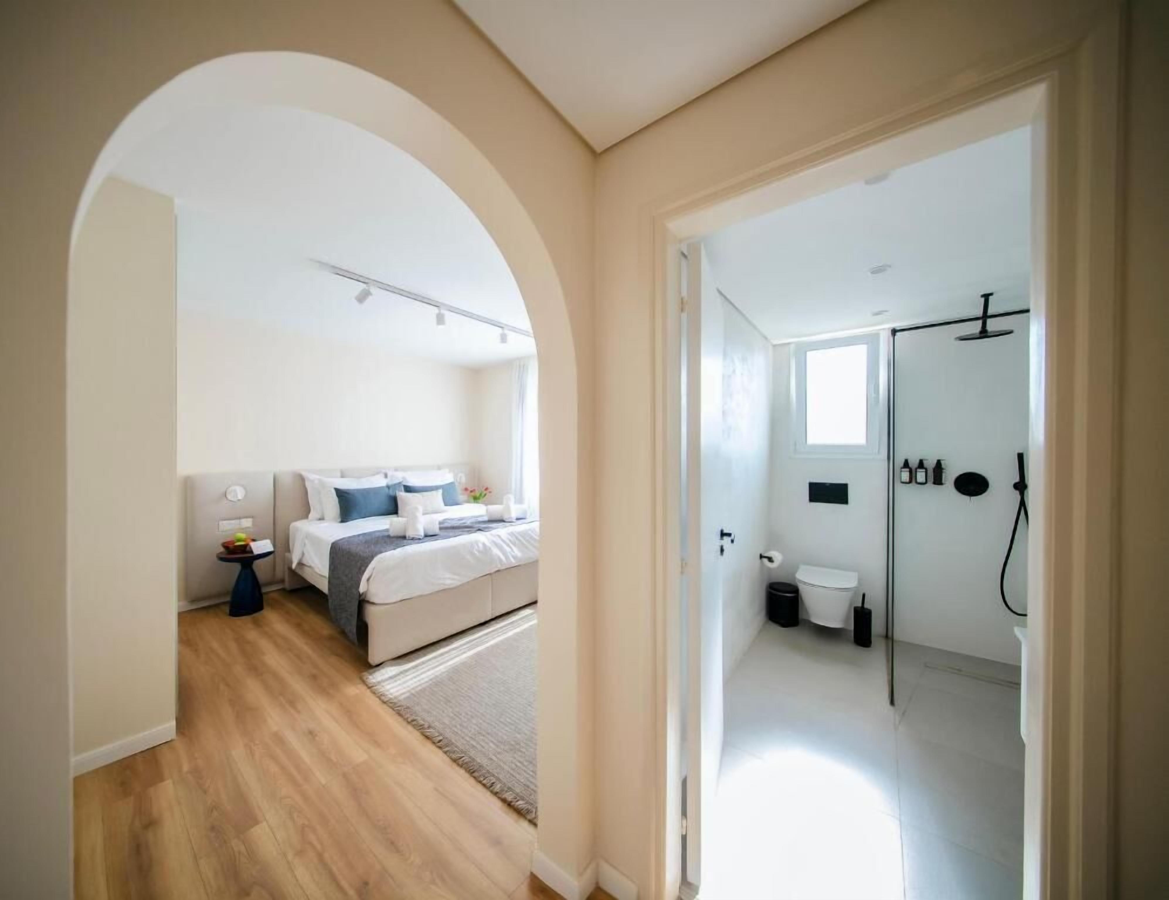 A bedroom with a bed, bedside table, and a view through an archway to a bathroom with a shower, toilet, and sink.