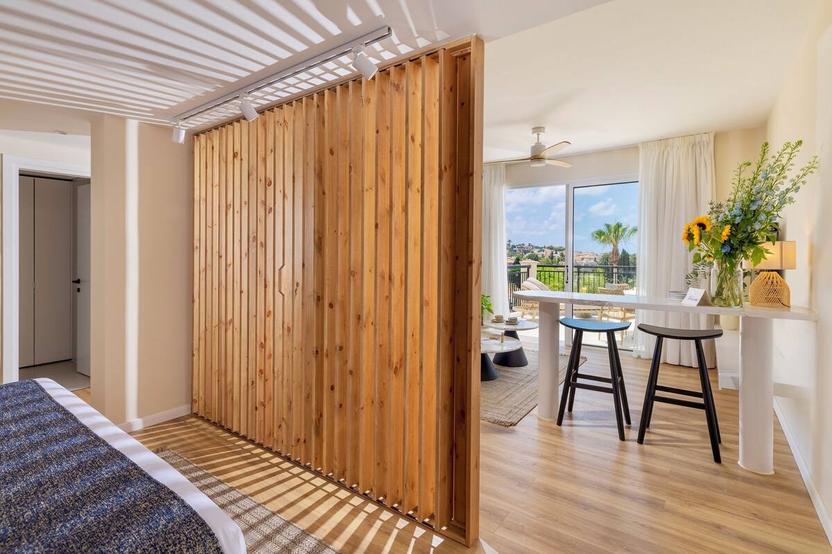 Quarto com cama, divisória de madeira e vista para uma varanda com mesa e cadeiras.