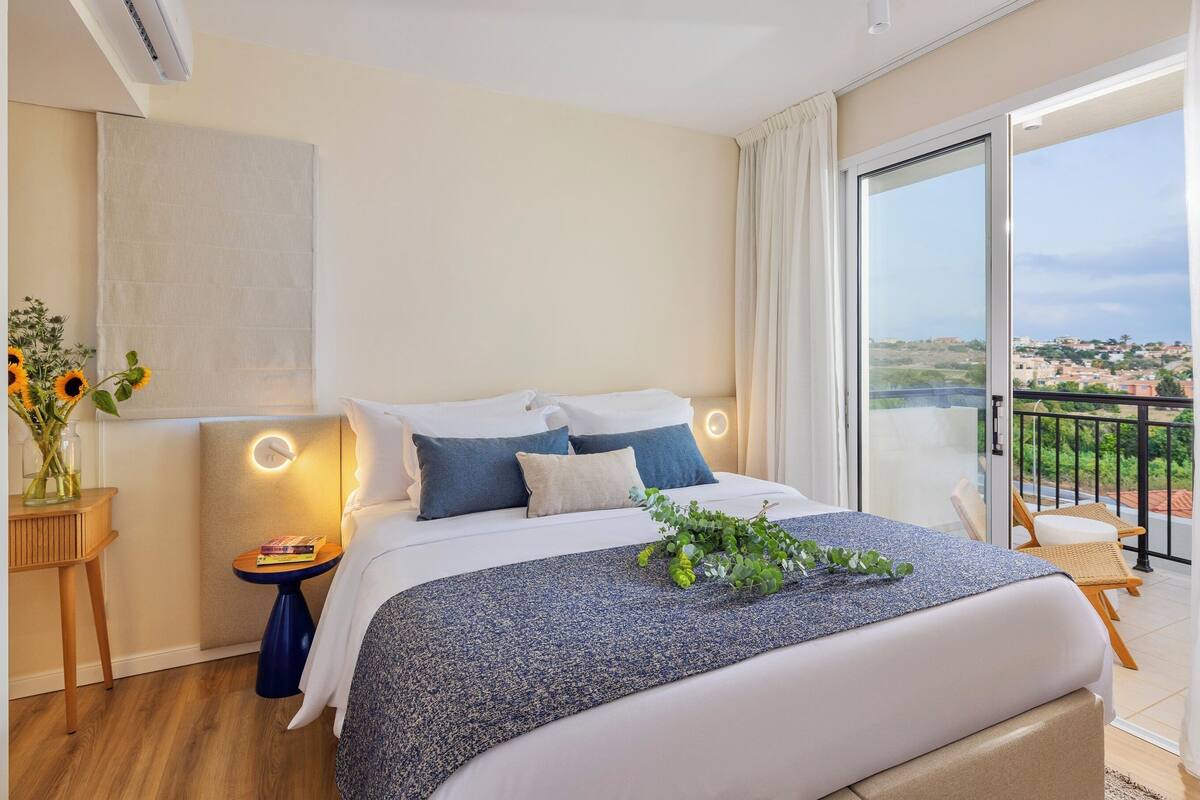 Quarto de hotel moderno com uma cama grande, uma mesinha de apoio, uma mesa de cabeceira, vista da cidade através de uma varanda e uma janela ampla com cortinas.