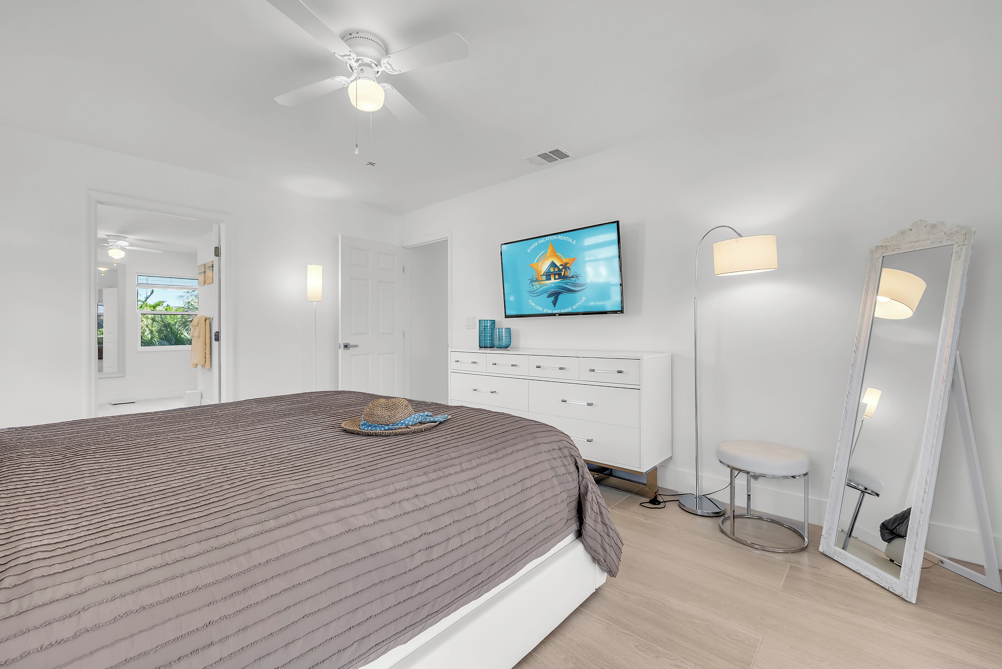 A bedroom with a bed, a ceiling fan, a TV, a dresser, a stool, and a mirror.