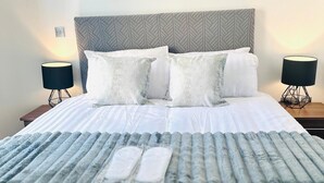 Una cama bien hecha con un edredón gris y blanco a rayas, dos almohadas blancas y un cabecero gris con un diseño geométrico. Hay dos mesitas de noche con lámparas negras a ambos lados de la cama.