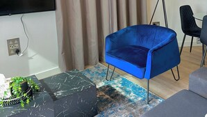 Una sala de estar moderna con un televisor de pantalla plana en la pared, un sillón de terciopelo azul, una mesa de centro de mármol negro con una planta decorativa, una lámpara de pie y un tapete estampado.