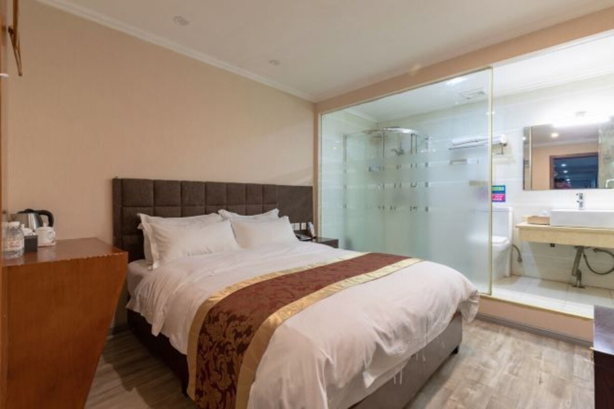 A hotel room with a bed, a wooden desk, a glass shower enclosure, and a bathroom with a sink and mirror.