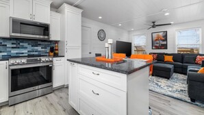 A modern kitchen with white cabinets, a stainless steel stove, and a black countertop. There is a dining area with a black sofa and orange cushions. - UNIQUE CONTAINER HOME/Branson Landing/Trolley Stop/Gazebo (Branson)