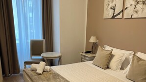 Habitación de hotel con una cama grande, una mesita de noche con lámpara, una silla y una ventana con cortinas.