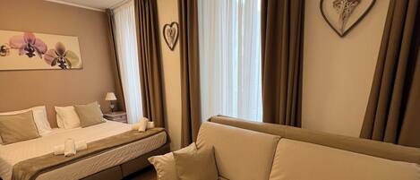 A hotel room with a bed, a sofa, wooden flooring, a painting, and curtains.