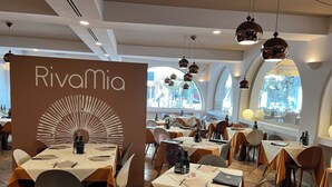 Interior de un restaurante con mesas dispuestas para comer, un llamativo letrero de 'Rivamia' y amplias ventanas que dejan entrar luz natural.