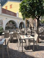 Un área de café al aire libre con mesas y sillas, un edificio amarillo con la señalización 'Rivamio' y un árbol que da sombra.
