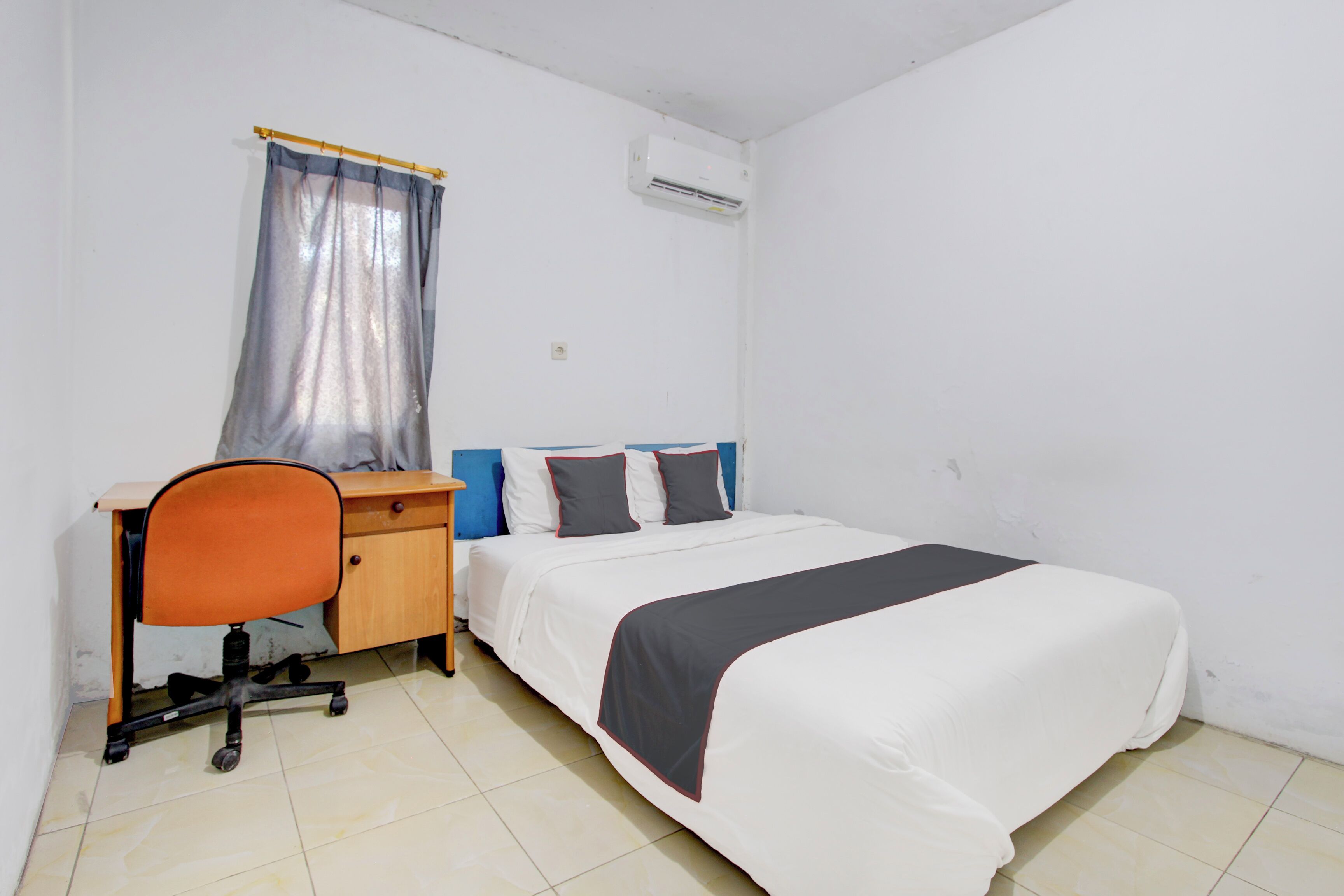 A bedroom with a bed, a desk, an office chair, a window with a curtain, and an air conditioning unit.
