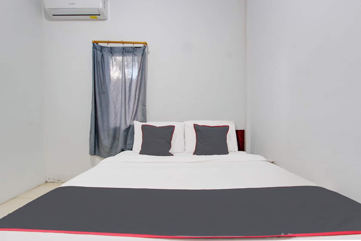 Una piccola camera con un letto singolo, copriletto bianco e nero, due cuscini, una testiera in legno, una finestra con tenda e un condizionatore d'aria a parete.