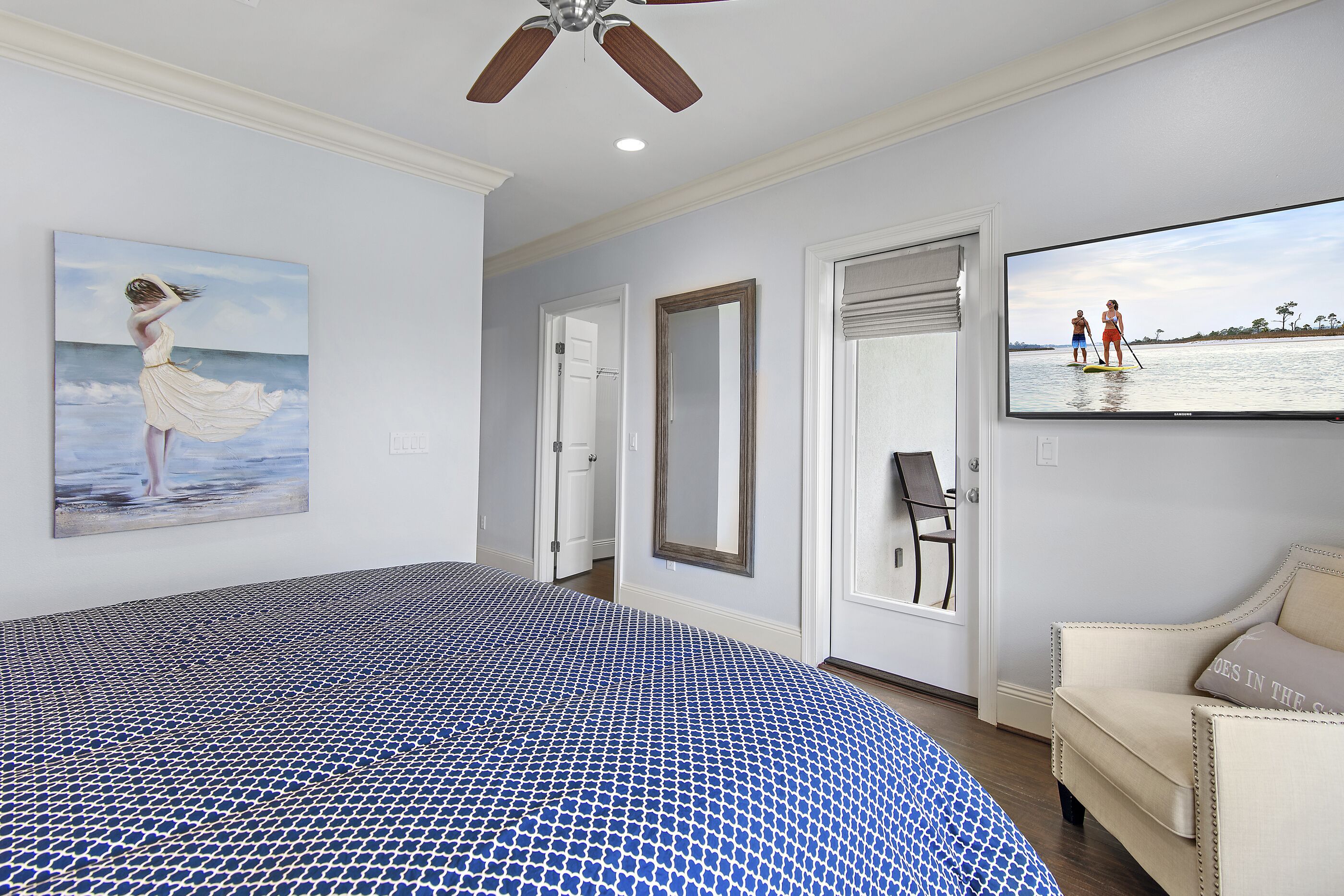 Ein Schlafzimmer mit einem großen Bett, einem Deckenventilator, einem Gemälde einer Frau am Meer, einem Fernseher und einer Tür zu einem anderen Raum.