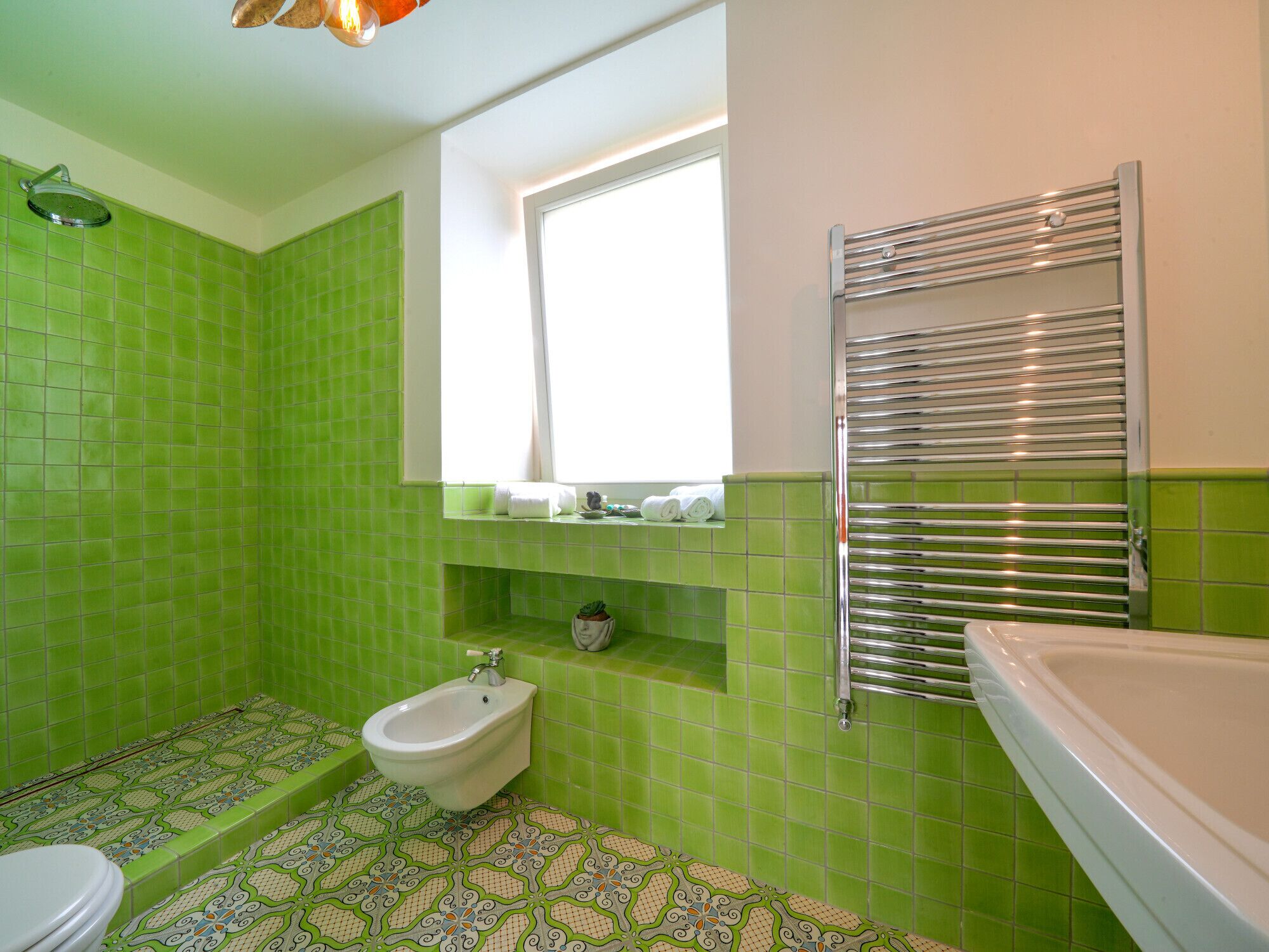 A bathroom with green tiles, a bidet, a window, and a towel rack.