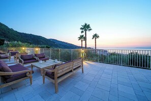 A terrace with outdoor furniture, a railing, and a view of the sea and hills.