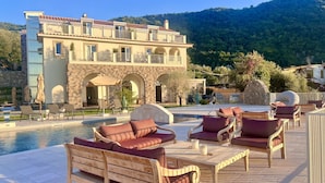 Zona junto a la piscina con sillones reclinables y una piscina, un edificio con fachada de piedra y balcones, y montañas al fondo.