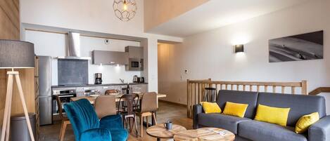 Una sala de estar moderna con un sofá gris, un sillón azul y una mesa de centro de madera. Se observa una cocina con electrodomésticos de acero inoxidable al fondo.