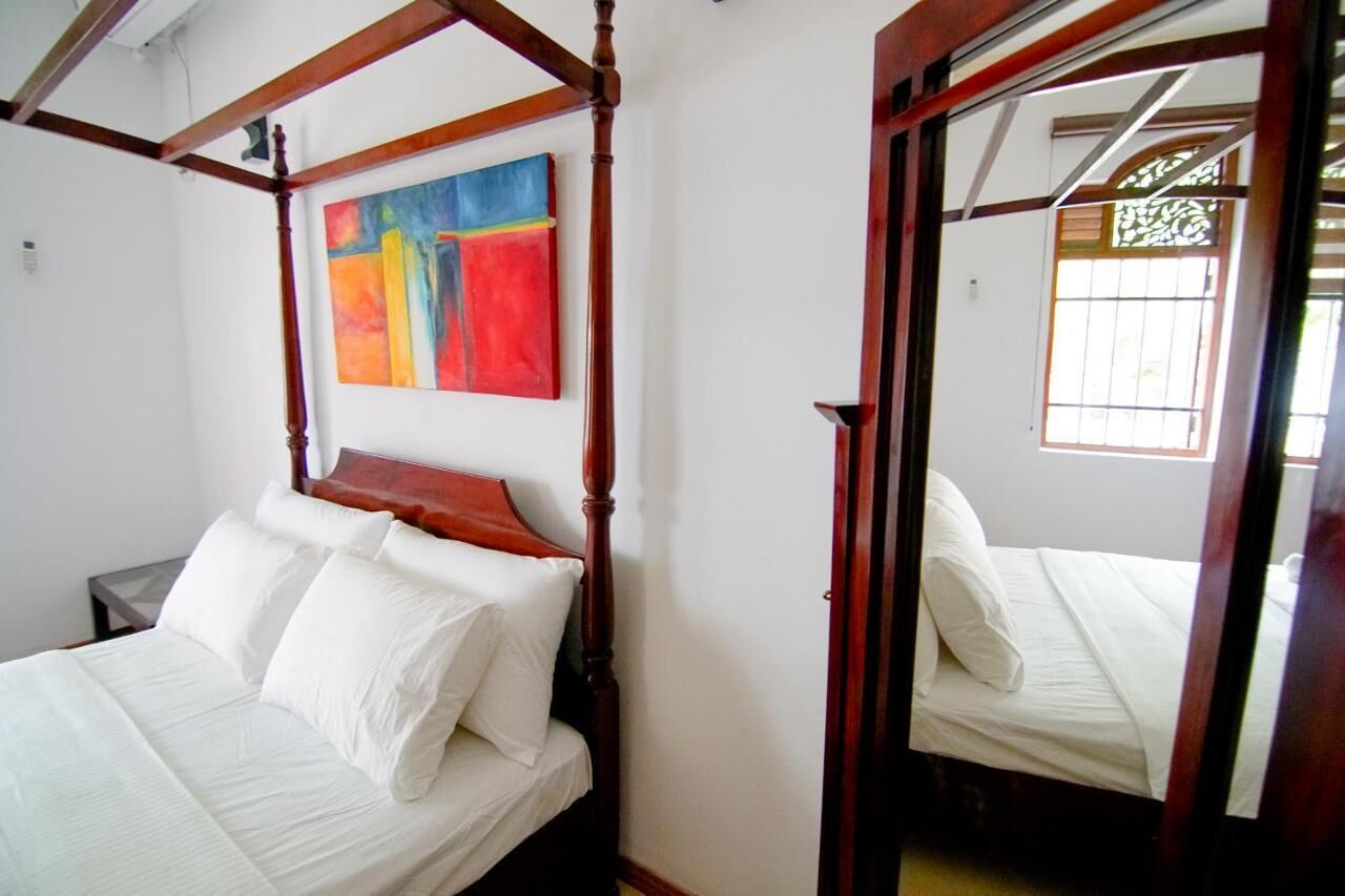 Un lit à baldaquin avec du linge de lit blanc, une peinture abstraite aux couleurs vives accrochée au mur et une fenêtre avec des barreaux.