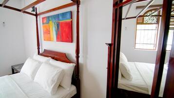 Un lit à baldaquin avec du linge de lit blanc, une peinture abstraite aux couleurs vives accrochée au mur et une fenêtre avec des barreaux.