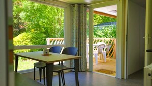 A room with a table and chairs, a view of greenery outside, and a wooden deck. - ZEcamping 4 étoiles piscine chiens acceptés club enfant mobil-home BBQ (La Bastide-de-Sérou)