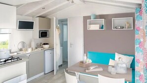 A compact kitchen with a gas stove, microwave, and a small refrigerator. There is a dining area with a table and chairs, and a teal-colored sofa. - ZEcamping 4 étoiles piscine chiens acceptés club enfant mobil-home BBQ (La Bastide-de-Sérou)