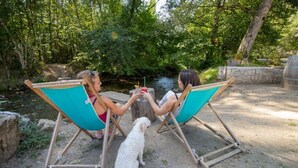 Two people sitting on turquoise chairs facing each other with a dog in between, near a river in a wooded area. - ZEcamping 4 étoiles piscine chiens acceptés club enfant mobil-home BBQ (La Bastide-de-Sérou)