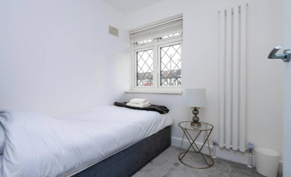 A small, white room with a single bed, a window with a view, a radiator, and a small table with a lamp.