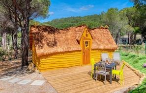 Une cabane en bois avec un toit de chaume, une terrasse avec des chaises et une vue sur une colline boisée.
