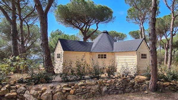 Une cabane en rondins, avec des fondations en pierre, entourée d’arbres et sous un ciel dégagé.
