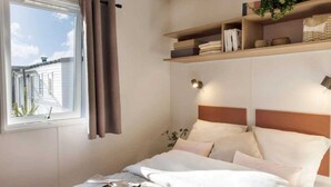A bedroom with a bed, a window with curtains, a shelf with books and decorative items, and a wall-mounted light. - ZEcamping 4* animé parc aquatique toboggans 500m plage locatif TV clim (Hyères)